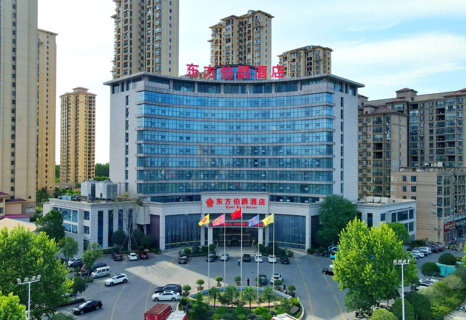 a large , modern building with a red and white sign in chinese characters , surrounded by other buildings and cars at Gushi Oriental Earl Hotel (Yucheng Avenue Genqin Culture Park)