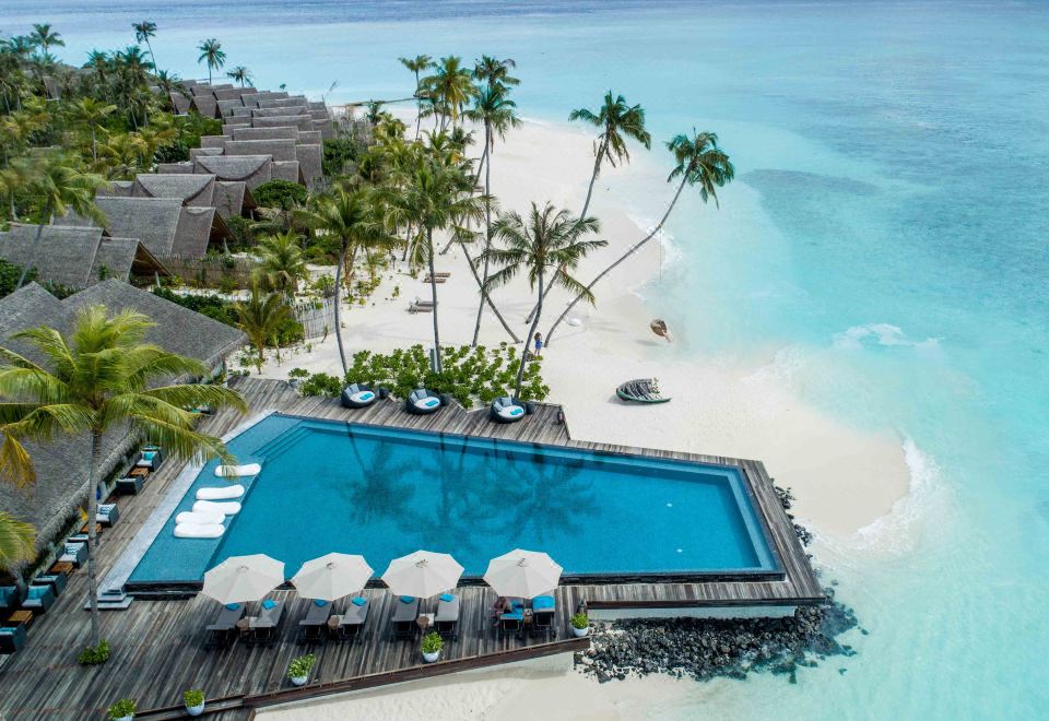 aerial view of a tropical beach with a large pool surrounded by palm trees , umbrellas , and lounge chairs at Fushifaru Maldives