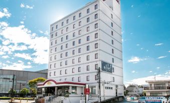 a white building with a red sign is situated on a street corner under a clear blue sky at Hotel Wing International Miyakonojo