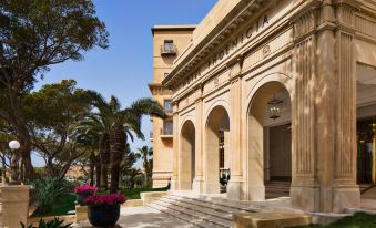 a grand stone building with columns and arches , surrounded by palm trees and pink flowers at The Phoenicia Malta