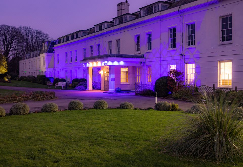 a large , white building with purple lights illuminating the entrance , surrounded by green grass and bushes at Avisford Park Hotel