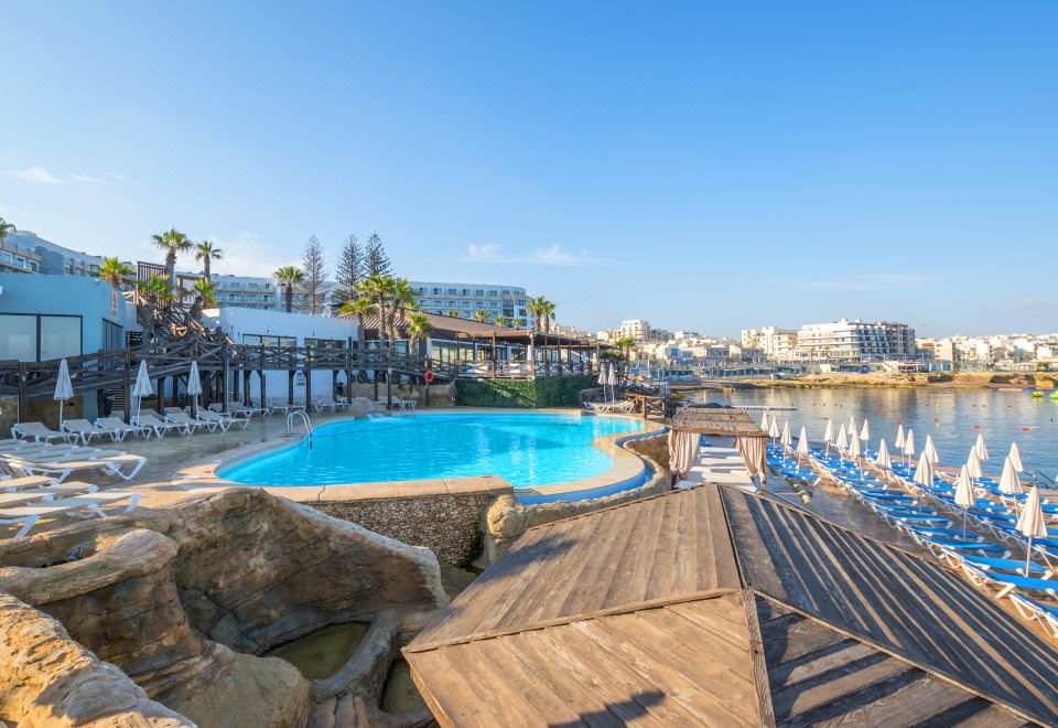 a large outdoor pool surrounded by lounge chairs and umbrellas , with a view of the water at DoubleTree by Hilton Malta