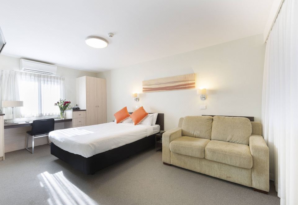 a large bed with orange pillows is in a room with a beige couch and windows at Ibis Styles Canberra Tall Trees