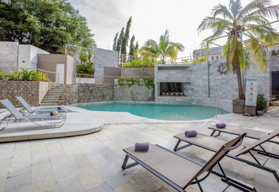 a beautiful outdoor area with a swimming pool , lounge chairs , and palm trees under a gray stone wall at Pearl Hotel