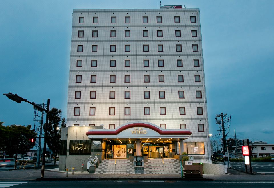 a large white building with a red roof and many windows is situated on a street corner at Hotel Wing International Miyakonojo