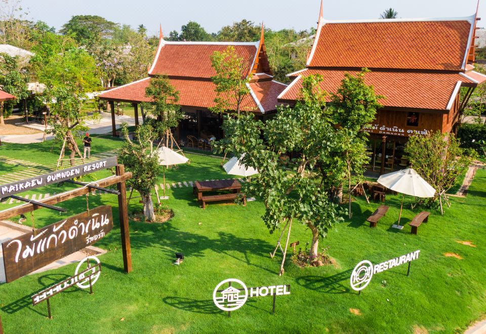 aerial view of a large red - roofed building surrounded by green grass and trees , with a restaurant nearby at Maikaew Damnoen Resort