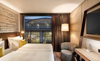 a hotel room with a bed , desk , chair , and large window offering a view of a modern building at HYPERION Hotel Leipzig