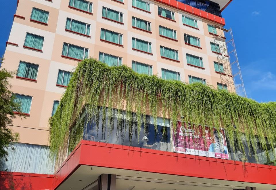 a large building with a red and white facade is surrounded by greenery , including a tree at Grand Zuri Dumai