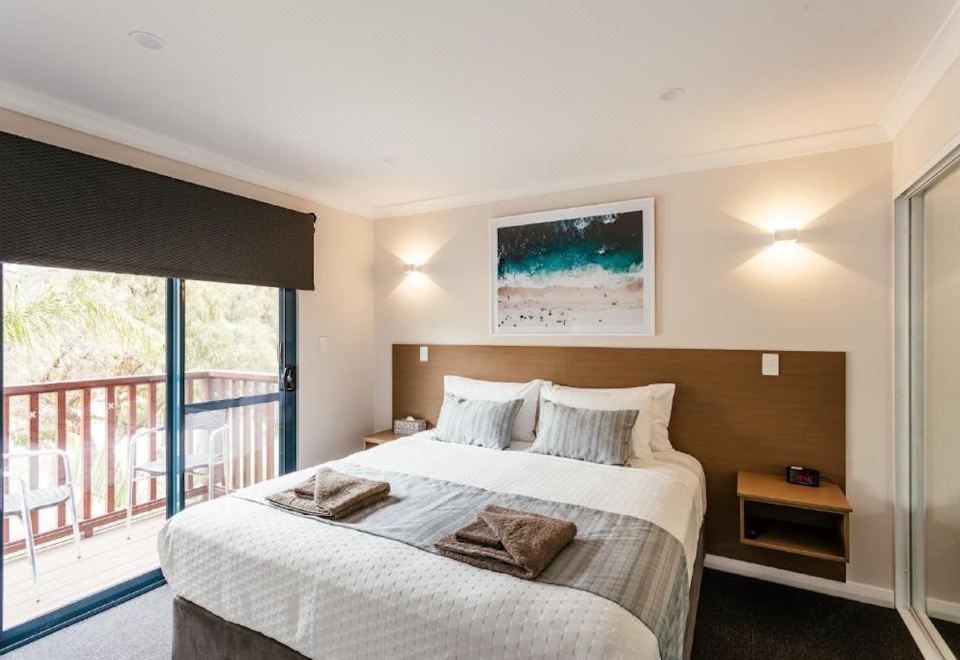 a large bed with white linens and a brown headboard is situated in a room with a window , lamps , and a painting at Dolphin Lodge Albany - Self Contained Apartments at Middleton Beach