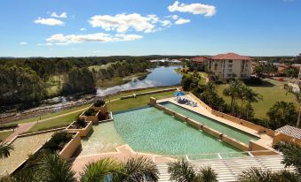 a large outdoor pool area with a water slide and lush greenery surrounding it , set against the backdrop of a river and buildings at Pelican Waters Resort