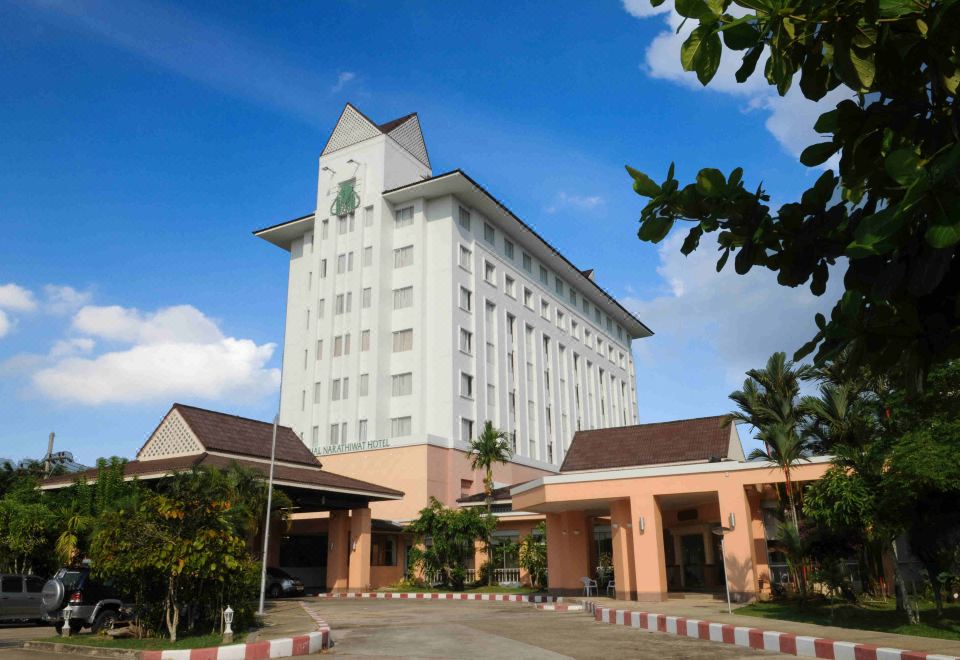 a large white building with a clock tower , surrounded by lush greenery and a clear blue sky at The Imperial Narathiwat Hotel