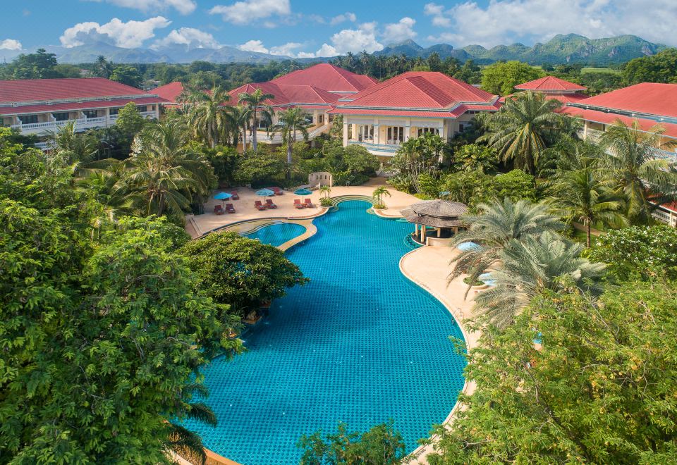 a large swimming pool is surrounded by lush greenery and palm trees in front of a red - roofed building at Dheva Mantra Resort