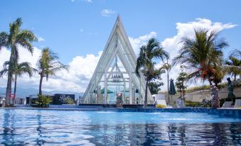 a modern glass structure with a triangular design is surrounded by palm trees and blue water at Swiss-Belhotel Silae Palu