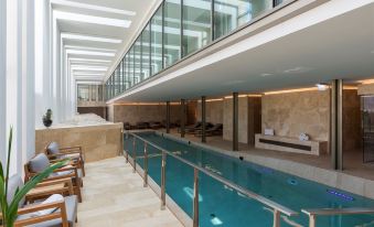 a large indoor swimming pool with a glass wall , surrounded by chairs and tables , in a modern building at The Phoenicia Malta