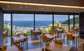 a modern restaurant with large windows offering a view of the ocean and cityscape , featuring wooden tables and chairs arranged for dining at Kamenoi Hotel Atami Annex