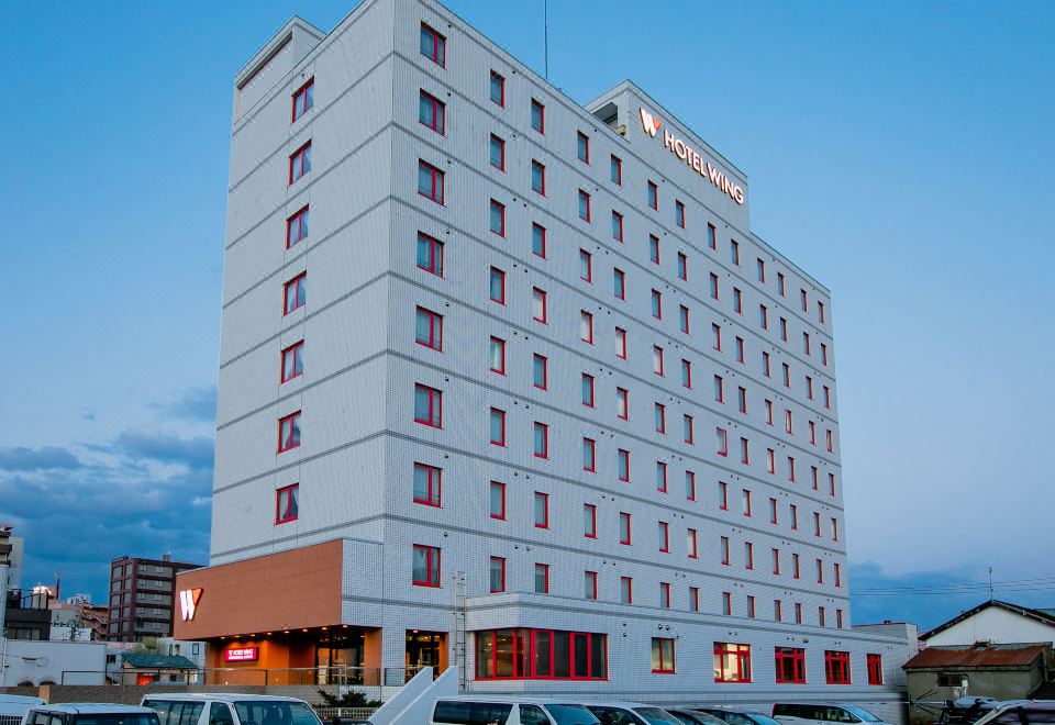 a tall white hotel building with a red roof , surrounded by a parking lot filled with cars at Hotel Wing International Chitose