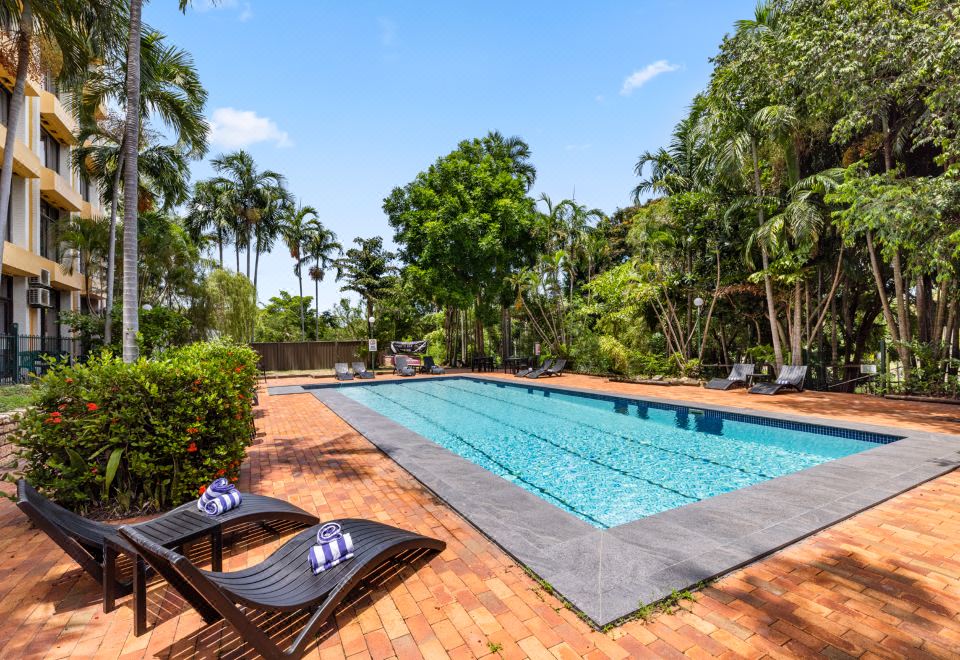 a large outdoor pool surrounded by lush green trees , with two lounge chairs placed near the edge of the pool at Frontier Hotel Darwin