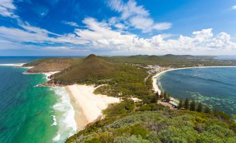 a panoramic view of a beach and coastline , with people walking along the shore and buildings scattered around the area at Oaks Port Stephens Pacific Blue Resort