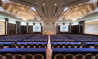 a large conference room with rows of blue tables and chairs , and two projectors on the wall at Hotel Metropolitan Akita