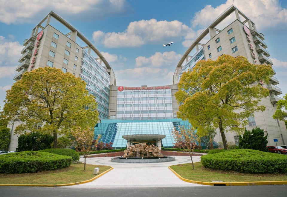 The picture shows a large building with an exterior view and its entrance, which is also part of the hotel at Ramada Plaza by Wyndham Shanghai Pudong Airport