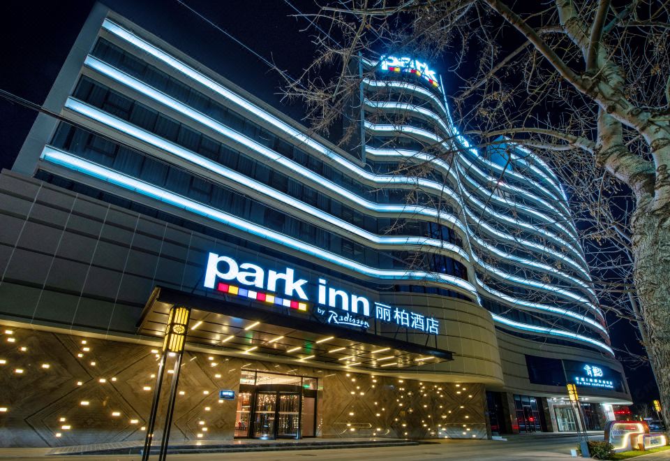 "At night, there is a front view of a building with a large sign on top that says ""hotel"" and other business names" at Park Inn by Radisson Beijing Tongzhou Universal Resort