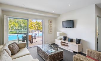 a modern living room with a flat screen tv , white furniture , and a sliding glass door leading to an outdoor pool area at Oaks Port Stephens Pacific Blue Resort