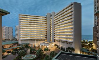 a large , modern building with multiple floors and balconies , illuminated at night , surrounded by palm trees and a pool at Hilton Myrtle Beach Resort
