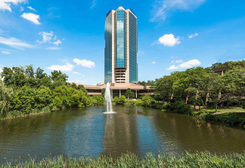 a tall building is reflected in a pond with a fountain , surrounded by trees and grass at Hilton Alexandria Mark Center
