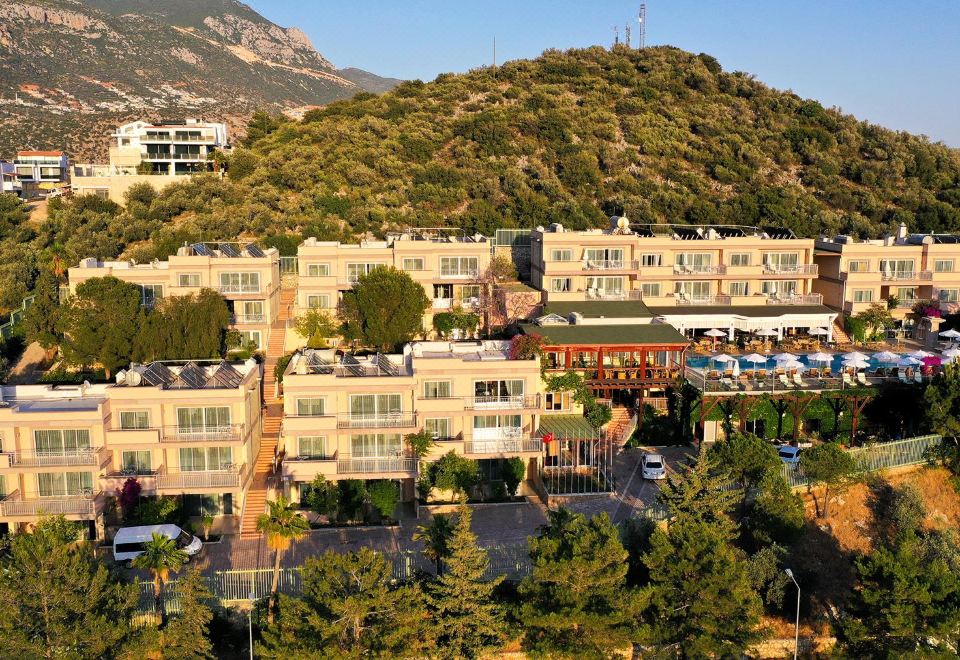 a residential area with multiple buildings , including a hotel , surrounded by trees and mountains , under a clear blue sky at Happy Hotel Kalkan