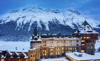 a large hotel surrounded by snow - covered mountains , with the sun setting in the background at Badrutt's Palace Hotel