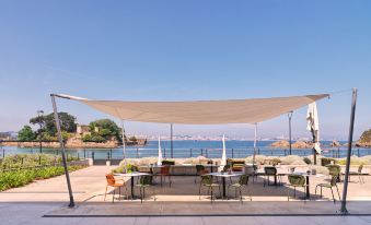 a sunny day at a beachside restaurant with umbrellas , tables , and chairs under a large white canopy , overlooking the ocean at Noa Boutique Hotel