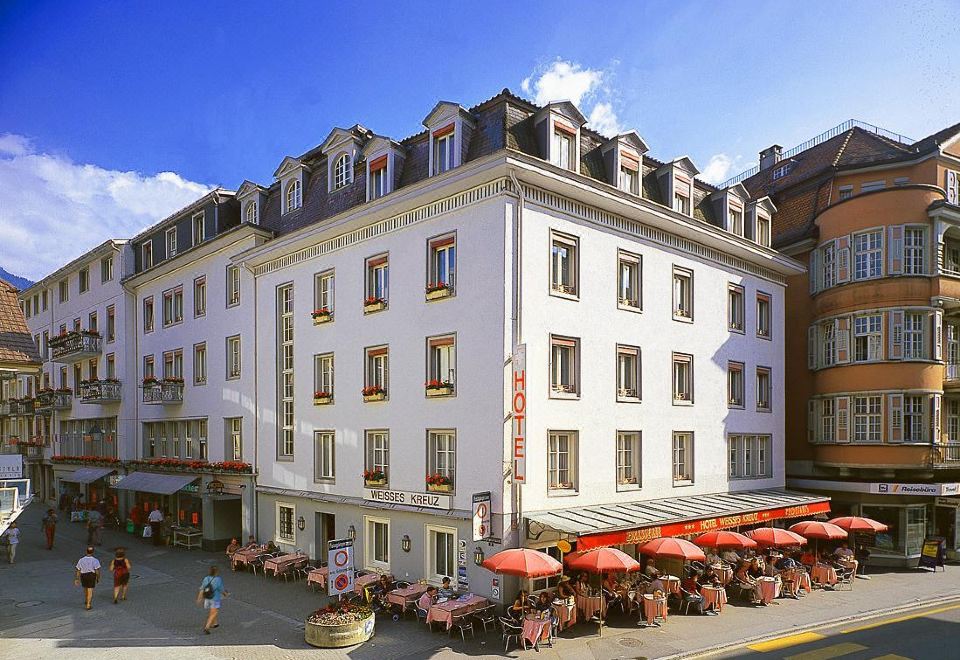 a large , white building with red umbrellas on a sunny day , surrounded by outdoor dining areas and people at Hotel Weisses Kreuz