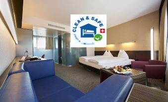 a hotel room with a bed , couch , and table is shown in the image , along with a clean & safe logo at Hotel Ambassador