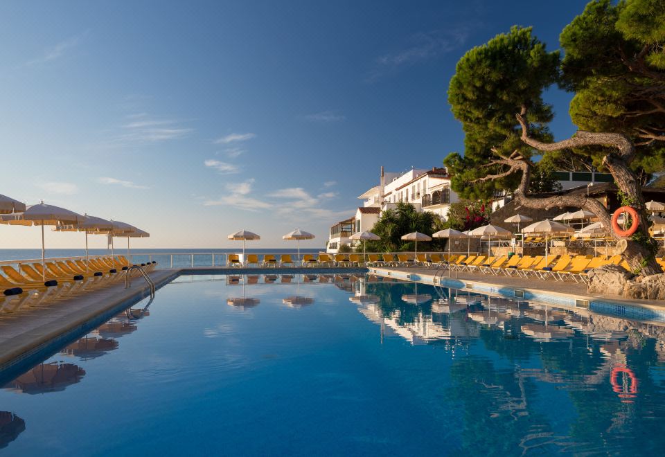 a large swimming pool surrounded by sun loungers and umbrellas , with a building in the background at Htop Caleta Palace #HtopBliss