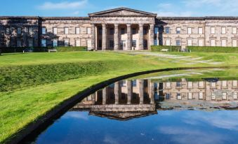 a large stone building with columns is reflected in a pool of water in a grassy area at Moxy Edinburgh Airport