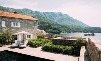 a stone building with a red roof , surrounded by greenery and mountains in the background at Aman Sveti Stefan