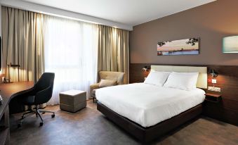 a large bed with white sheets is in a room with a chair , ottoman , and window at Hampton by Hilton Toulouse Airport