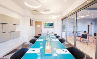 a long conference table with blue chairs , glasses , and papers is set up in a room with large windows at Holiday Inn Express Stockport