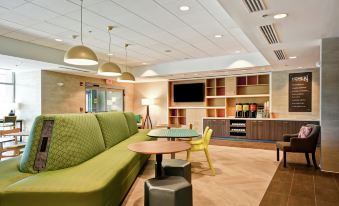 a modern office lobby with a green couch , wooden tables , and hanging lights , as well as a television and shelves at Home2 Suites by Hilton Walpole Foxboro