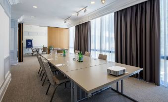a conference room with a long table and chairs , water bottles , and a window with brown curtains at NH Luxembourg