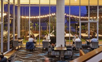 a group of people sitting at a table in a restaurant with large windows and outdoor seating at Mercure Melbourne Caroline Springs