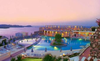 a large outdoor swimming pool surrounded by multiple buildings , with the sun setting in the background at Athina Palace Resort & Spa