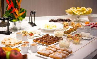 a table is filled with various desserts , including donuts , cakes , and fruit , arranged in a visually appealing manner at Simon Hotel