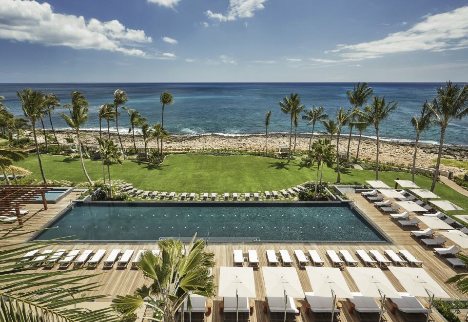 a large pool surrounded by lounge chairs and palm trees , with the ocean visible in the background at Four Seasons Resort Oahu at Ko Olina
