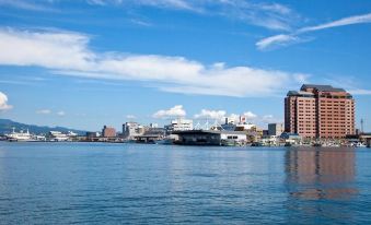 a city skyline with buildings and trees , set against a blue sky with white clouds , while the water reflects the reflection of the buildings at La Vista Hakodate Bay