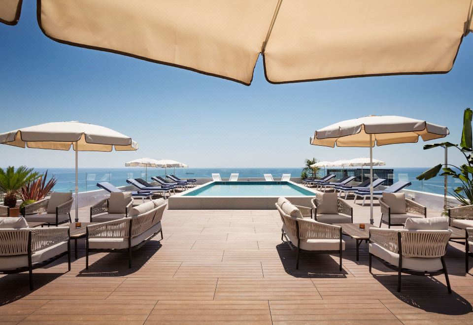 a large swimming pool is surrounded by lounge chairs and umbrellas , with the ocean visible in the background at H10 Imperial Tarraco