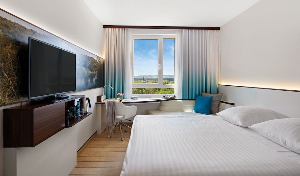 a modern hotel room with a large window , white bed , and blue curtains , as well as a desk , chair , tv , and other at Hey Lou Hotel Frankfurt Airport
