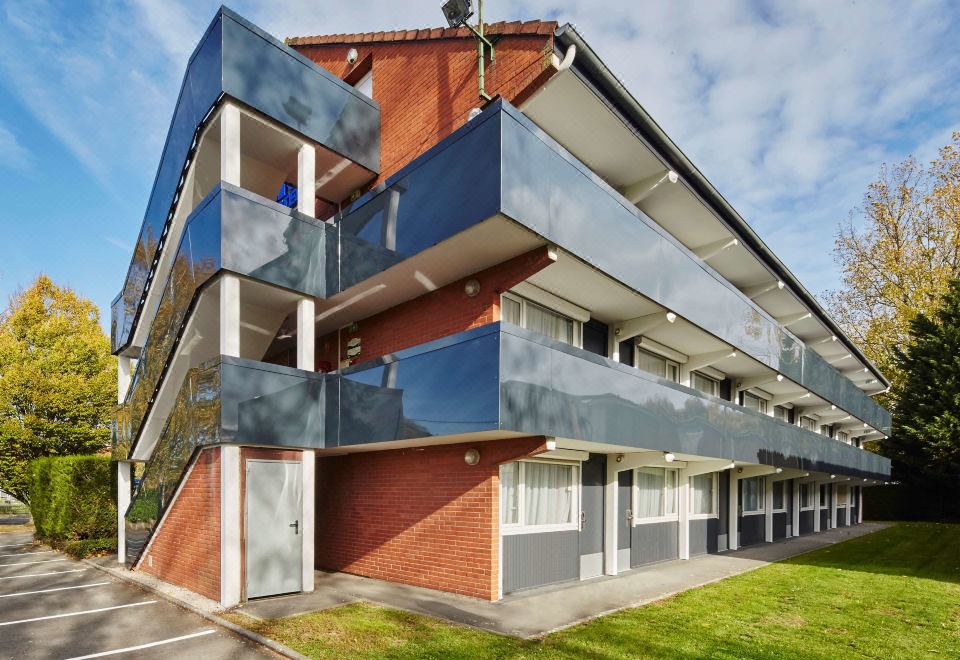 a modern apartment building with a red brick exterior and blue accents is surrounded by green grass at Kyriad Lille Est - Hem