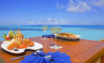 a wooden dining table with two wine glasses and a plate of food placed on it , overlooking the ocean at Medhufushi Island Resort
