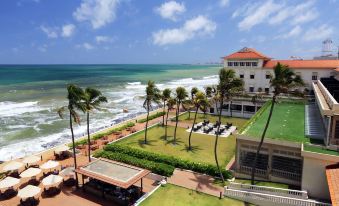 a beach scene with a large white building and palm trees in the background , overlooking the ocean at Galle Face Hotel
